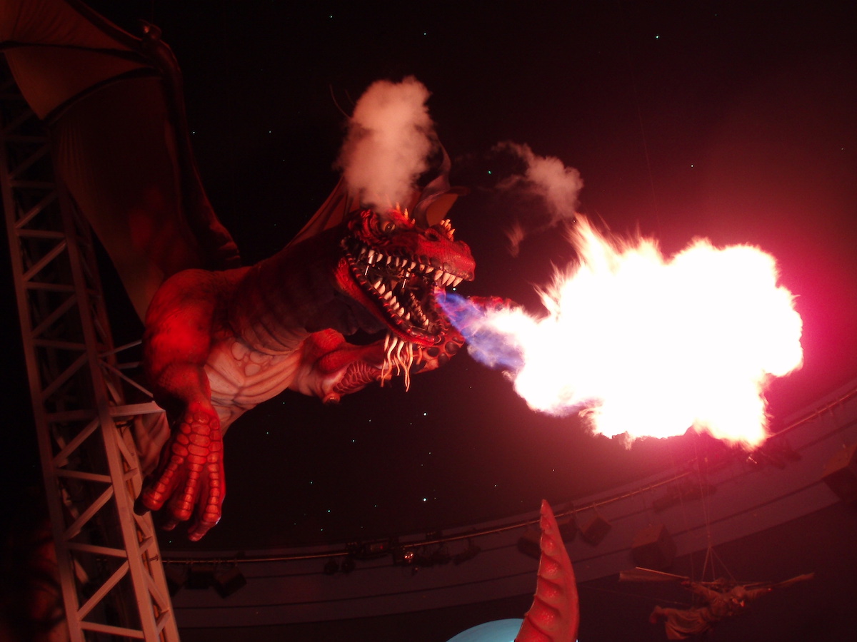 A large animatronic dragon exhales a plume of flames against a darkened red background in an indoor space.