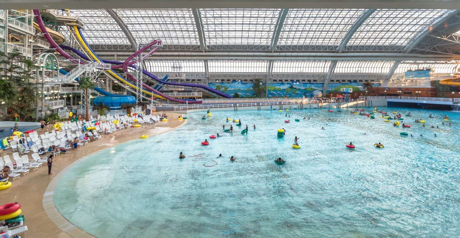 A view of a large indoor pool features people floating on colourful inner tubes and, to the left of the frame, white lawn chairs set up against a fake 'beach' with the floor a sandy colour. Networks of long, colourful tubular waterslides tangle in the upper left quadrant of the frame.