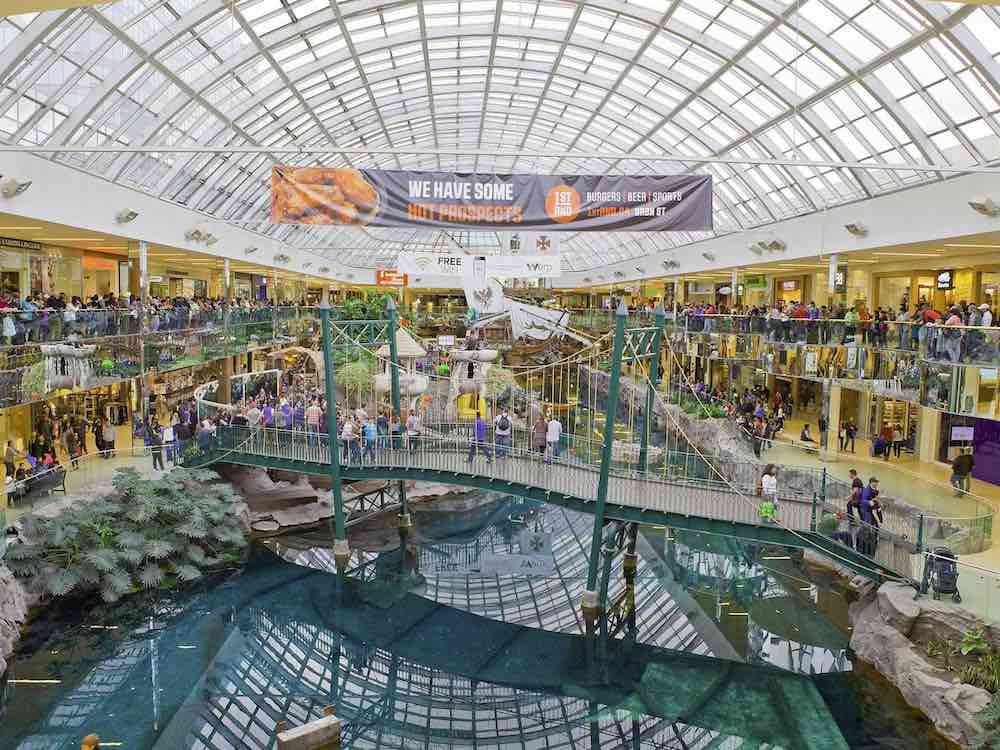A shopping mall&rsquo;s galleria features a rounded ceiling with white scaffolding over an aquamarine waterway running through the middle of the promenade, flanked by shops and walkways on either side. The waterway features fake plants and rocks, and a small green cable bridge features a pedestrian walkway for shopping mall patrons.
