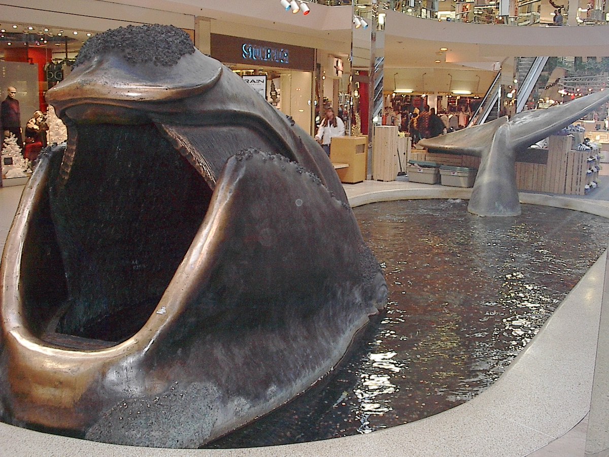 A large grey metallic sculpture of an open-mouthed whale stands in an oblong water feature in the galleria of a shopping mall.
