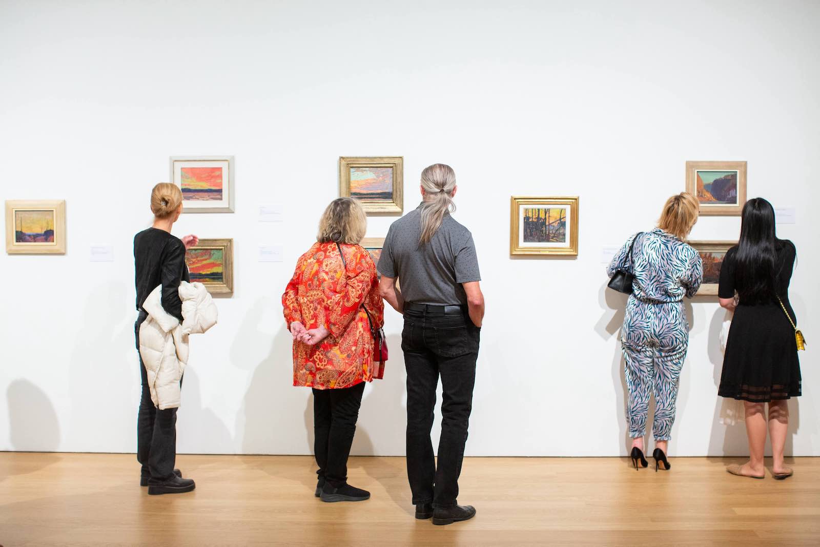 Five people stand with their backs to the camera as they look at small framed oil paintings mounted across a wide white gallery wall. They are dressed in contemporary clothing and standing on a light hardwood floor.
