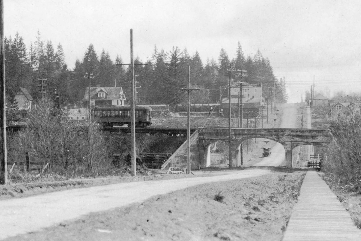 An old black and white photo of the interurban tracks on a bridge over dusty Boundary Road. A train is crossing the bridge; an old car approaches the underside of the bridge.