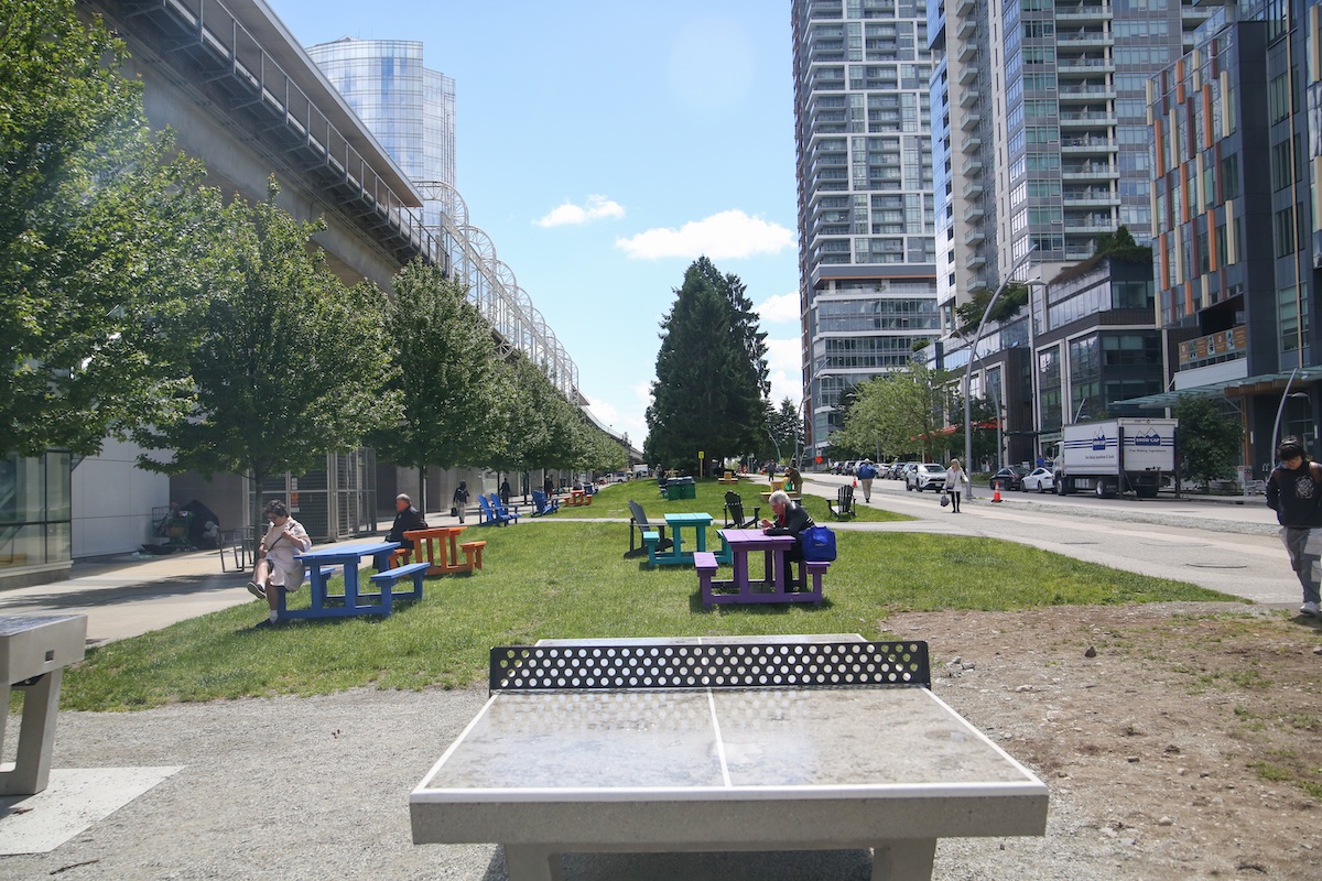 A permanent, concrete ping-pong table with picnic tables in the background, to the right of the elevated SkyTrain tracks.