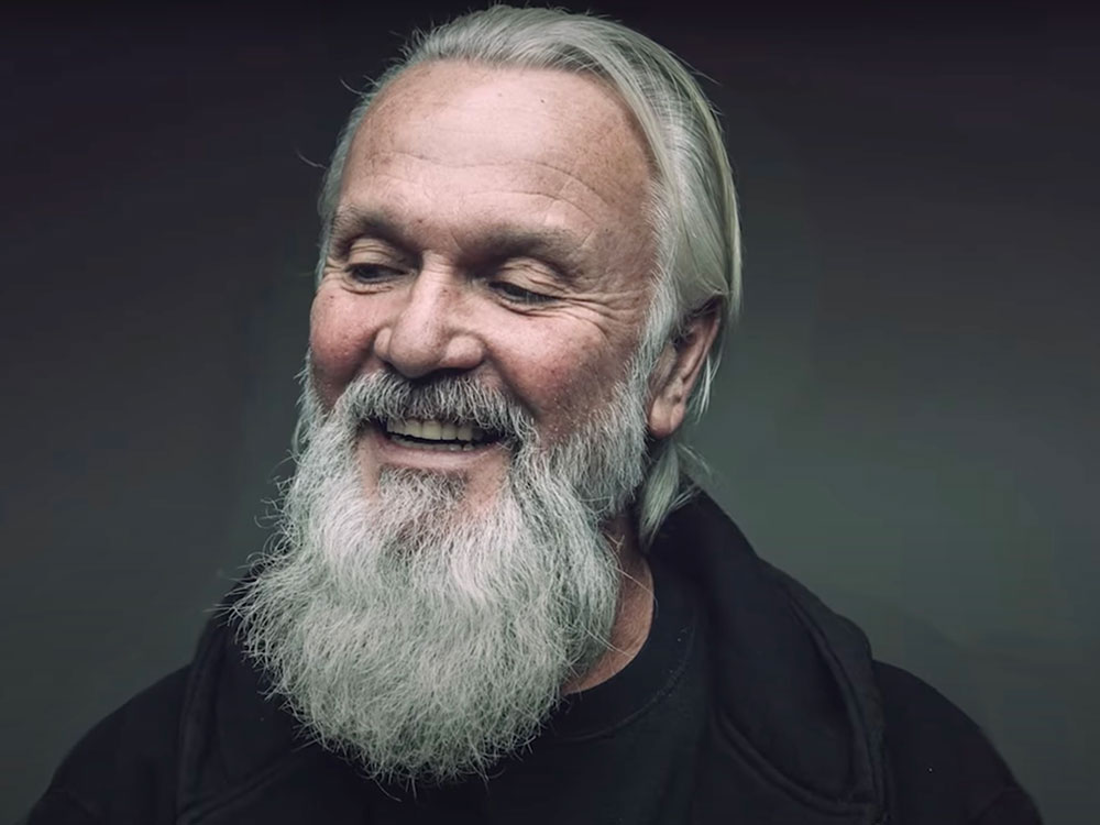 Barry Shantz has long grey hair and a bushy grey beard. He wears black and is looking down towards the left of the frame, smiling. He is seated against a grey background.