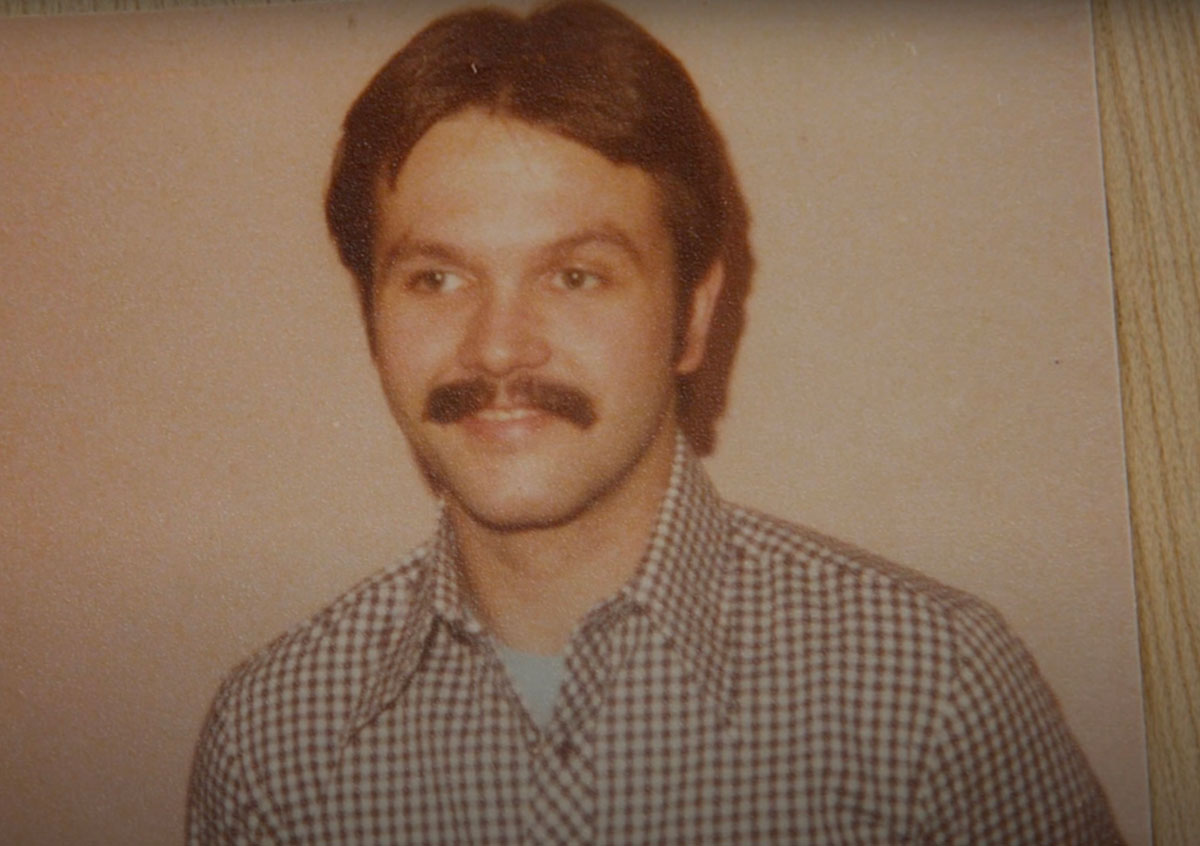 An archival photo of Barry Shantz portrays a young man with short dark hair and a moustache in a brown gingham button-up shirt. He looks to the left of the frame, smiling softly.