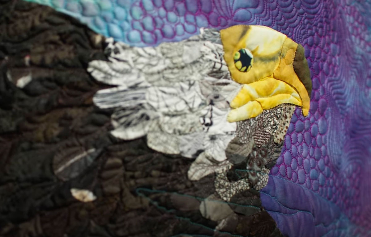A close-up of the quilt shows an eagle, done in appliqu&eacute; style, on a textured purple background. The style the artist has chosen for the eagle&rsquo;s body mimics feathers in quilt form. 