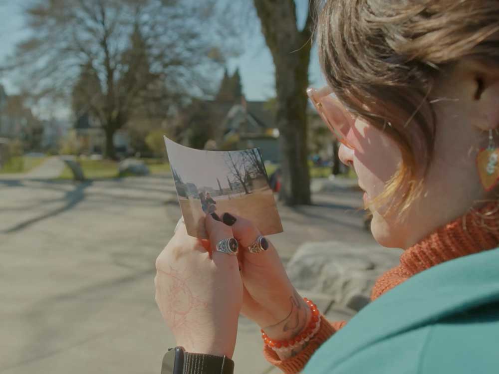 jaye simpson has short, wavy, light brown hair. She is wearing rose-coloured sunglasses and a turquoise jacket and standing in a sunny urban park. The camera is behind her shoulder while she holds an old photograph of an adult holding a young child. Her nails are painted black.
