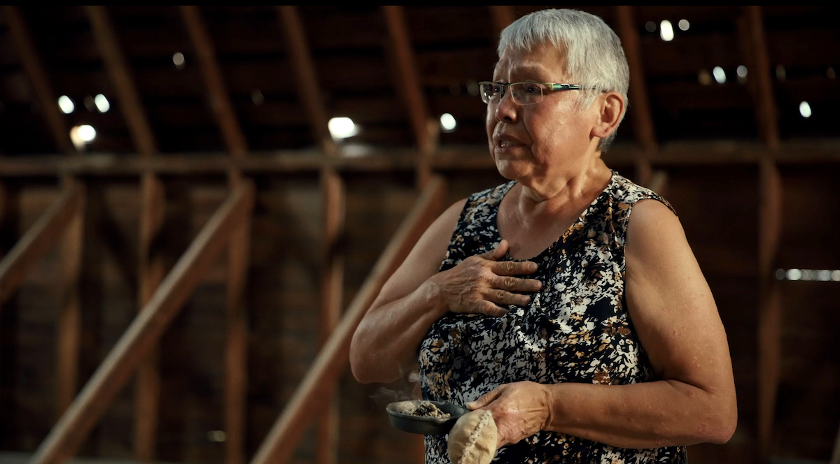 An older woman with medium skin tone and very short grey hair holds a small cast iron pan in her left hand and holds her right hand over her heart. She is visibly emotional.