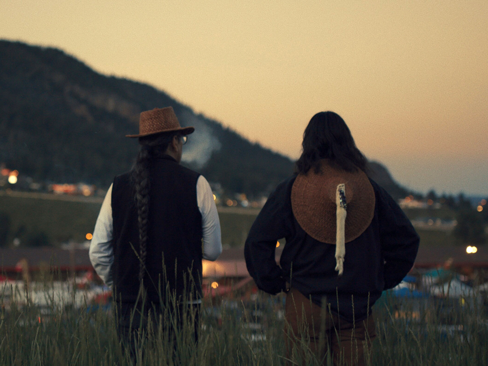 Two men with long black hair stand with their backs to the camera. One wears a woven hat and the other has a similar hat hanging from his neck. It is dusk. In the distance there are lights and mountains beyond.