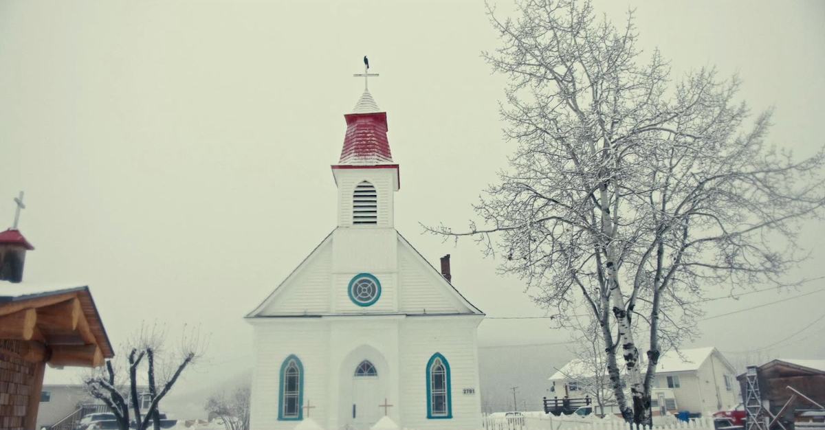 The facade of a small white clapboard church amidst the snow. Part of the roof near the spire is red.