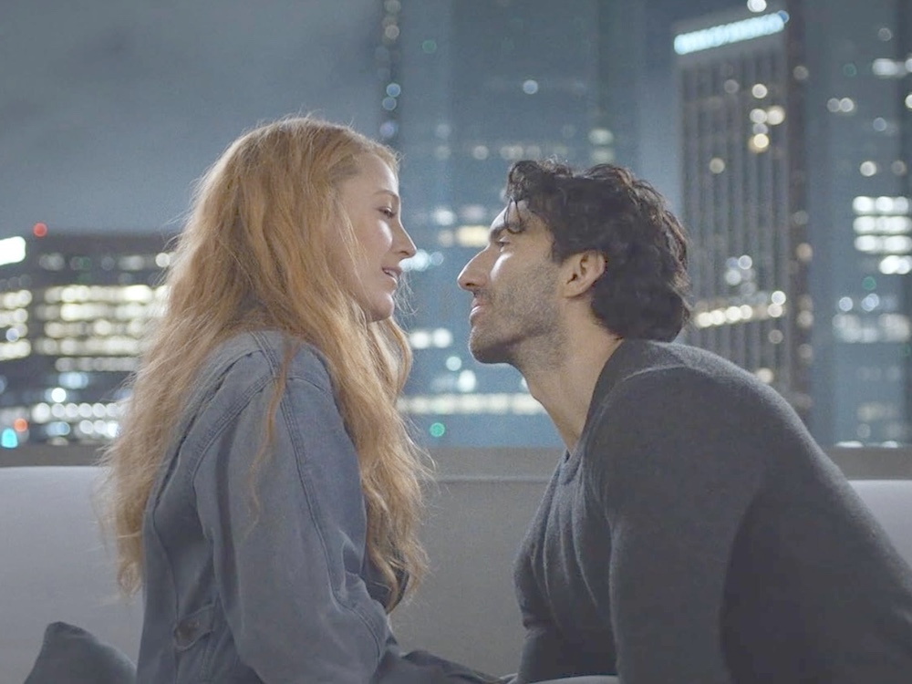 Actress Blake Lively is a white woman with long wavy blond hair. She is seated on a sofa in a downtown apartment at night next to Justin Baldoni, a white man with short dark wavy hair. They are leaning towards each other as if they are about to kiss.