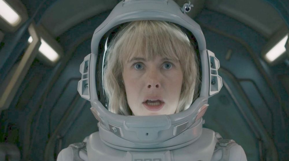 A white woman with blonde hair and light skin is in a space suit, looking to the horizon with an expression of shock. She is seated in a darkened spacecraft.