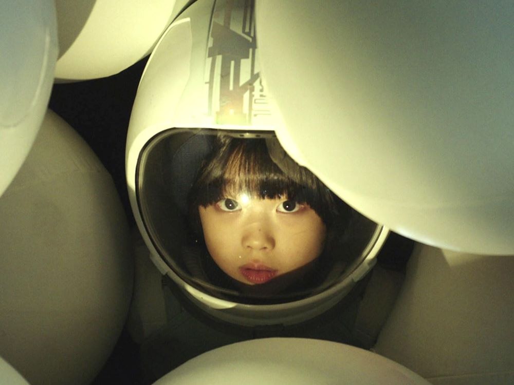 A Korean child in a white astronaut’s helmet looks up from a mass of white spherical objects in a dimly-lit space.