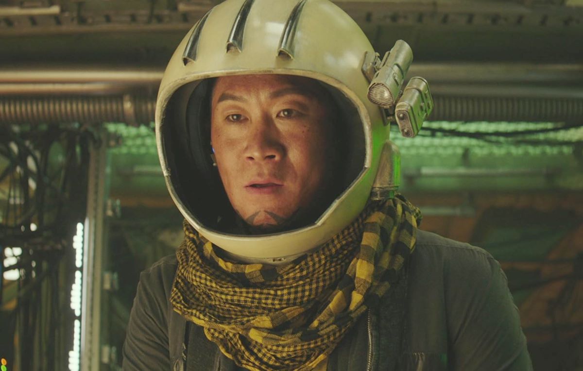 A Korean man wears a white astronaut’s helmet, a black and yellow keffiyeh scarf and a dark green hoodie in a dimly lit industrial-style space.