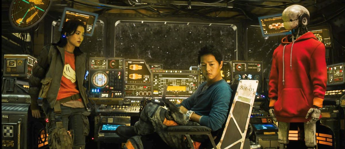 Two actors and a robot clad in a red hoodie stand in costume on the bridge of a spacecraft alight with antiquated-looking control panels. A woman stands on the left in casual streetwear over a red T-shirt; a man is seated on a metal chair in a blue pullover with a pack on his lap.