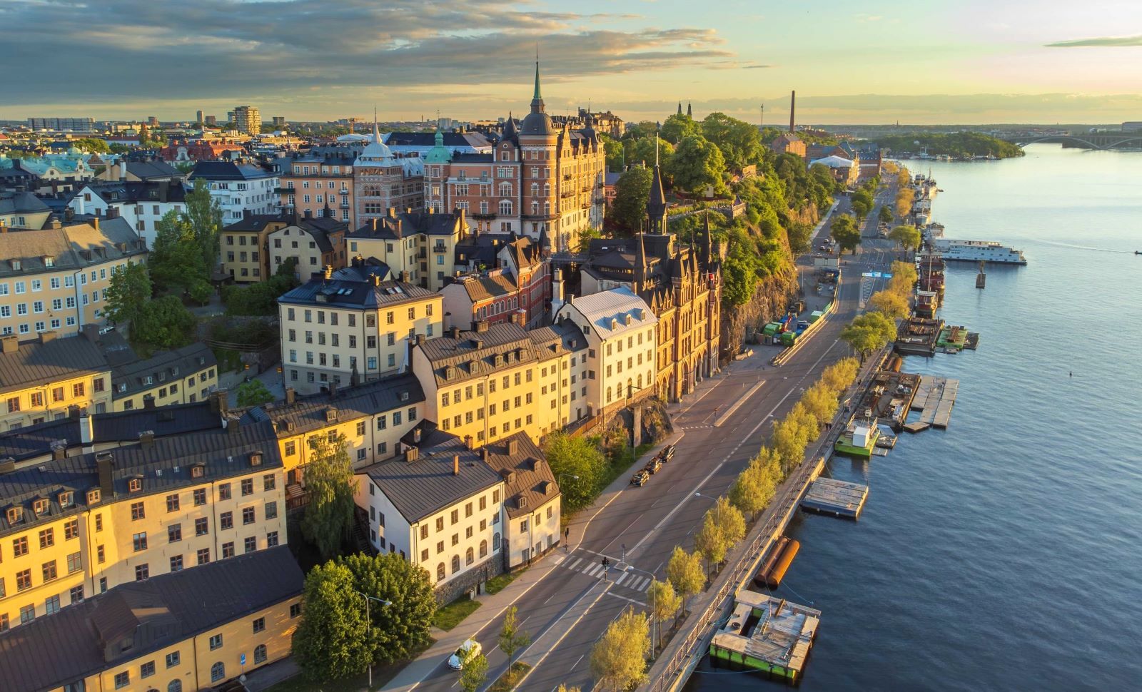 A view of Sweden&rsquo;s S&ouml;dermalm district at sunset casts heritage buildings in a golden light. To the right of the frame is a road, docks and water.