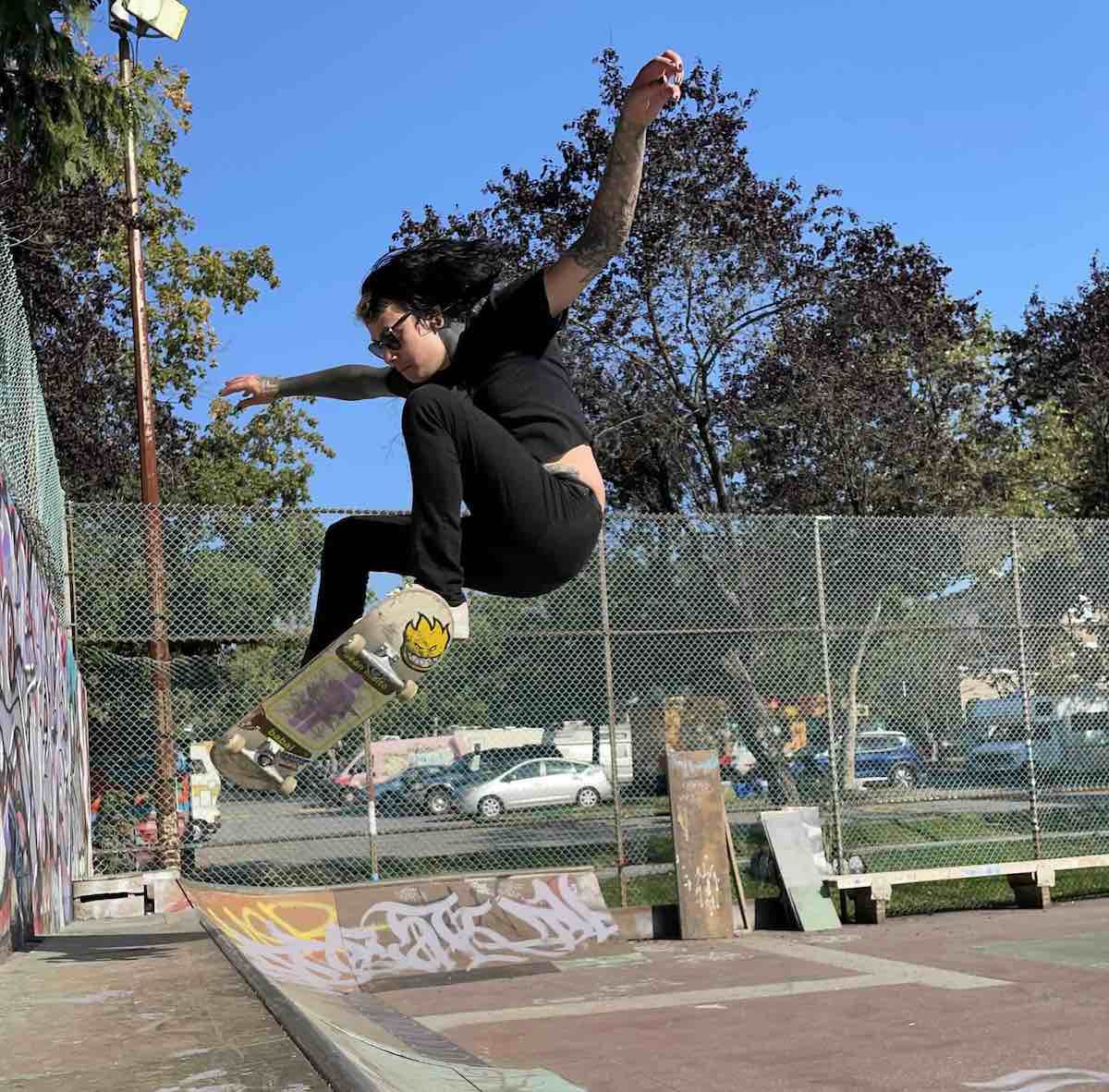 Dakota Cootes has long dark hair and is wearing sunglasses, a black T-shirt and black pants. She is in the midst of a jump on her skateboard above the top of a ramp. Behind her is a tall chain-link fence, a stand of trees on a residential street. The sky is blue and it’s a sunny day.
