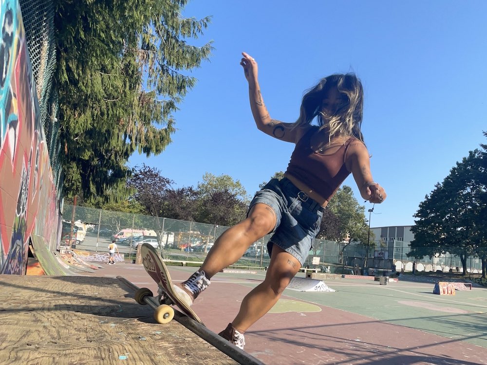 Kim Regala’s dark wavy hair obscures her face as she rides a skateboard up a ramp in a spacious outdoor skate park on a sunny day. She is wearing summer clothing: a maroon tank top, jean shorts and maroon canvas high-top sneakers.