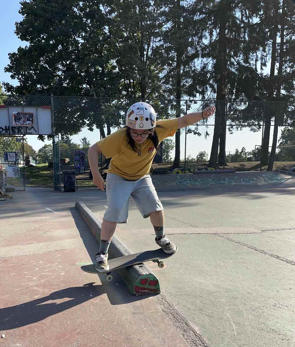 Li Charmaine Anne navigates her skateboard over a concrete barrier. She is wearing a white helmet decorated with stickers, a yellow T-shirt and jean shorts. She has long dark hair and glasses, and behind her is a stand of trees behind a chain-link fence on a bright, sunny day.