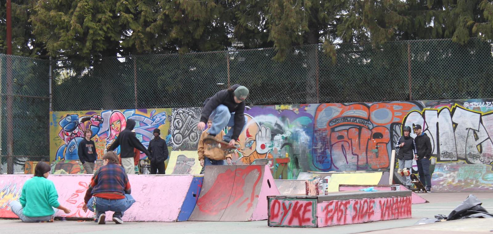 A skateboarder in a green toque and black hoodie on a skateboard, jumping over a high ramp. Around and behind them are other skaters gathered and socializing or repairing nearby ramps.