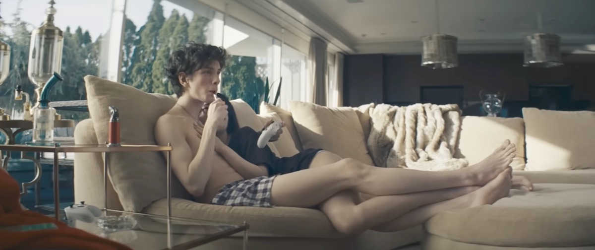 Mark Eydelshteyn wears navy and white plaid boxers and stretches out his legs to rest across a beige sofa. He holds the stick of a lollipop to his mouth in one hand and a white video game console in the other. Actor Mikey Madison is curled up next to him.