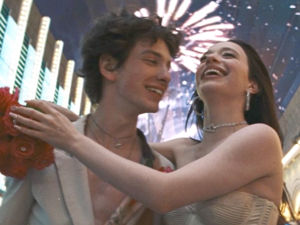A young man and woman have their arms around each other, smiling widely. Actor Mark Eydelshteyn, left, has short dark curly hair and light skin and is wearing a cream-coloured blazer over a bare chest. Actor Mikey Madison, right, has long dark hair and pale skin and is wearing a strapless cream-coloured bustier with sparkling jewelry. She holds a bouquet of fake red roses. Fireworks are in the sky in the background.