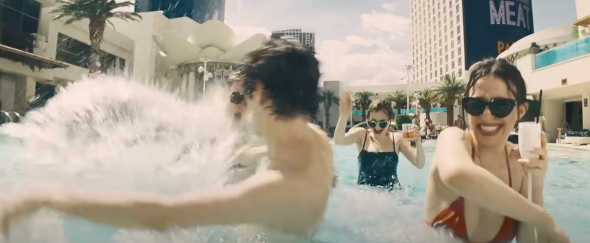 Young adults splash around in an outdoor pool flanked by highrises. On the left, actor Mark Eydelshteyn has his back turned to the camera as he splashes in the water. On the right, actor Mikey Madison holds a white cup in one hand and raises her elbow up, smiling. She’s wearing sunglasses and a red bikini.
