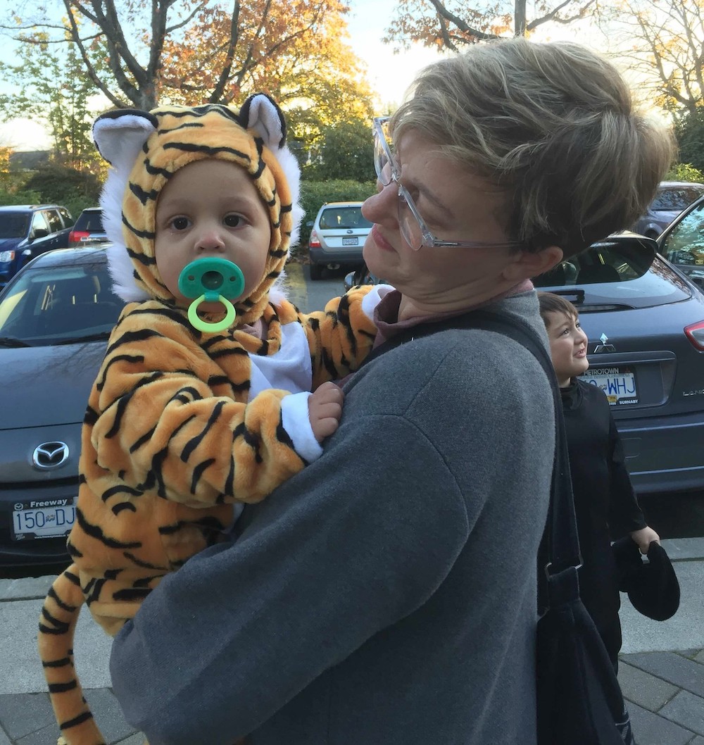 A woman in a grey sweatshirt with short blond hair and glasses holds a toddler in a furry orange and black tiger costume with a green pacifier in her mouth.