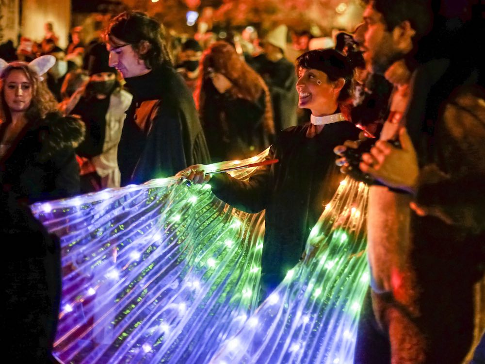 A woman with dark hair in an updo holds a brightly lit pair of rainbow butterfly wings. To her left, a man wears an animal onesie while holding a small child on his shoulders. They are among a crowd of costumed people on a street at night.
