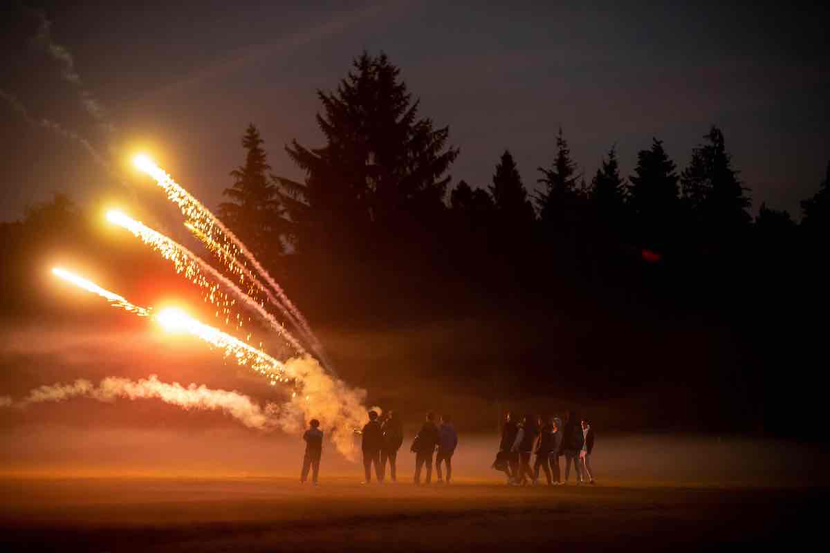 Silhouettes of young men are situated on an open field against a shadowy orange background lit by the lights of fireworks. Behind them is a dark tall stand of coniferous trees.