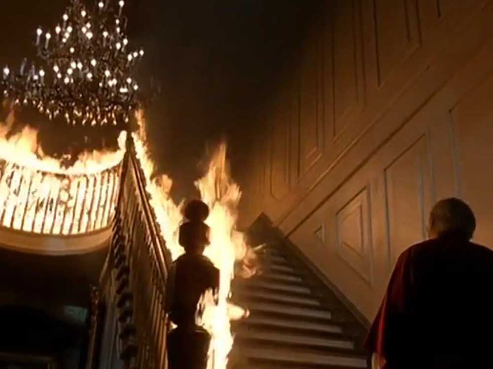 A man in a robe stands to the right of the frame with his back turned to the camera. He is at the bottom of a large indoor staircase under a chandelier. The railings are on fire.
