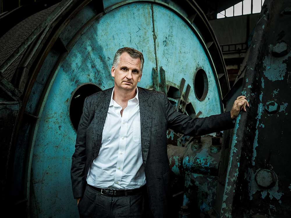 Timothy Snyder has short greying hair and light skin. He wears a dark grey suit and a white button-down shirt with no tie. He is standing with one hand resting on a rusted piece of railing. Behind him is a large, rusting circular piece of industrial equipment painted turquoise.