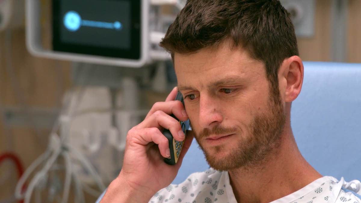 Brett has short brown hair, light skin and a short beard. He is seated in a hospital bed and wearing a white patterned hospital gown. He holds a black phone to his ear and is looking down.