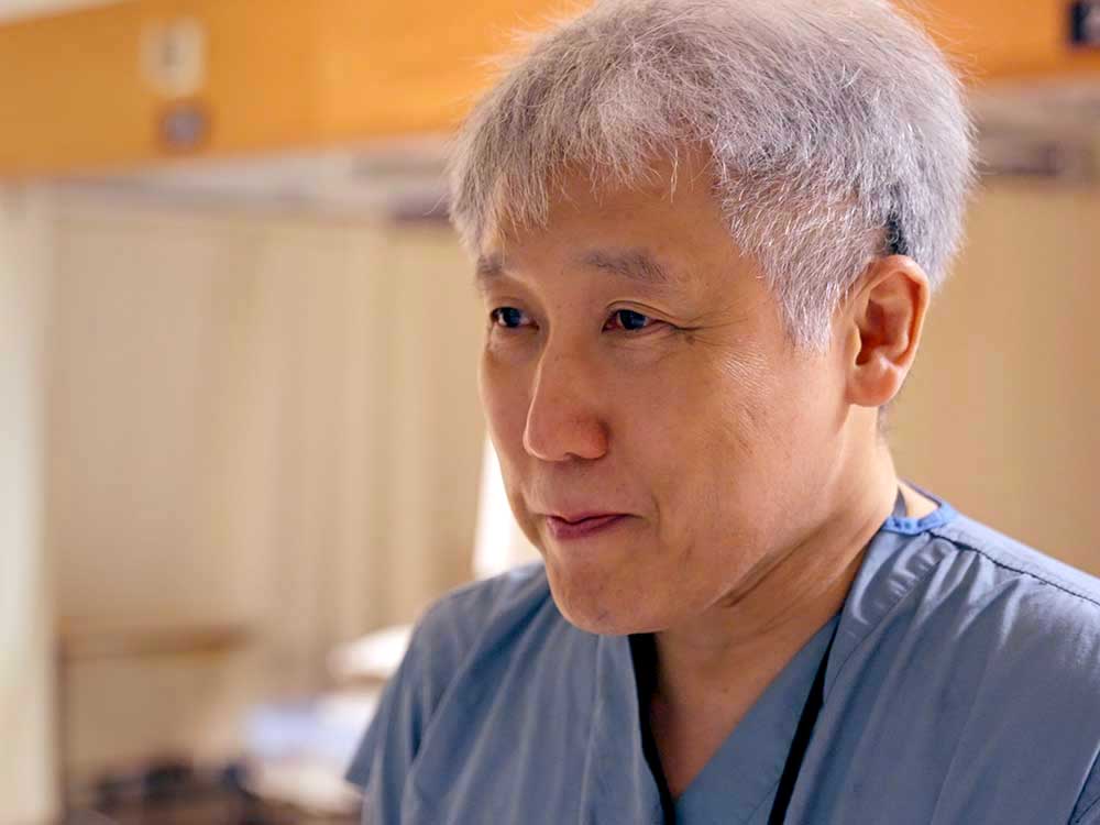 Dr. John Yee has short grey hair and medium skin. He is wearing blue medical scrubs and looking towards the left of the frame with a tender expression. Behind him in soft focus is the interior of a hospital.