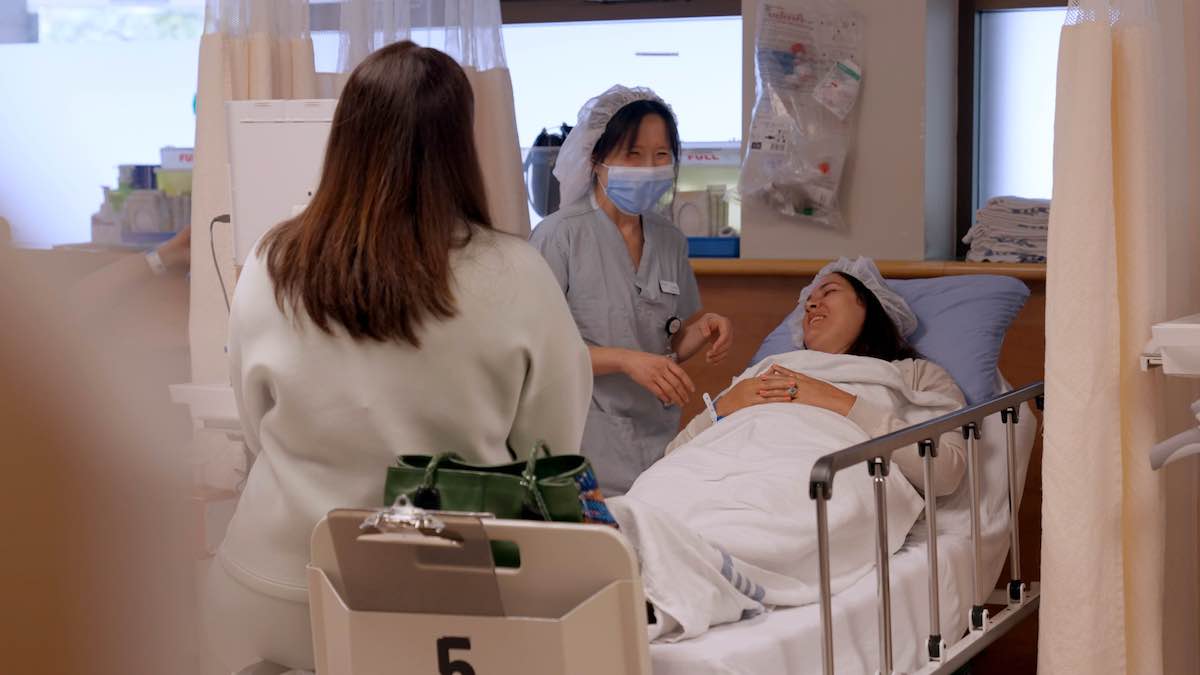 Marcela has long dark hair and tanned skin and is wearing a hairnet. She is wrapped in white hospital blankets and is smiling alongside two people at her bedside, one of whom has their back turned to the camera. The person facing the camera has black hair, blue scrubs, a hairnet and a blue medical mask.