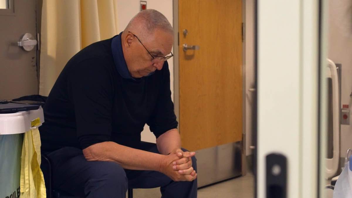 A man with buzzed grey hair, glasses, light skin and a dark button-down shirt and jeans sits on a chair in a hospital corridor. His hands are clasped and he looks down.