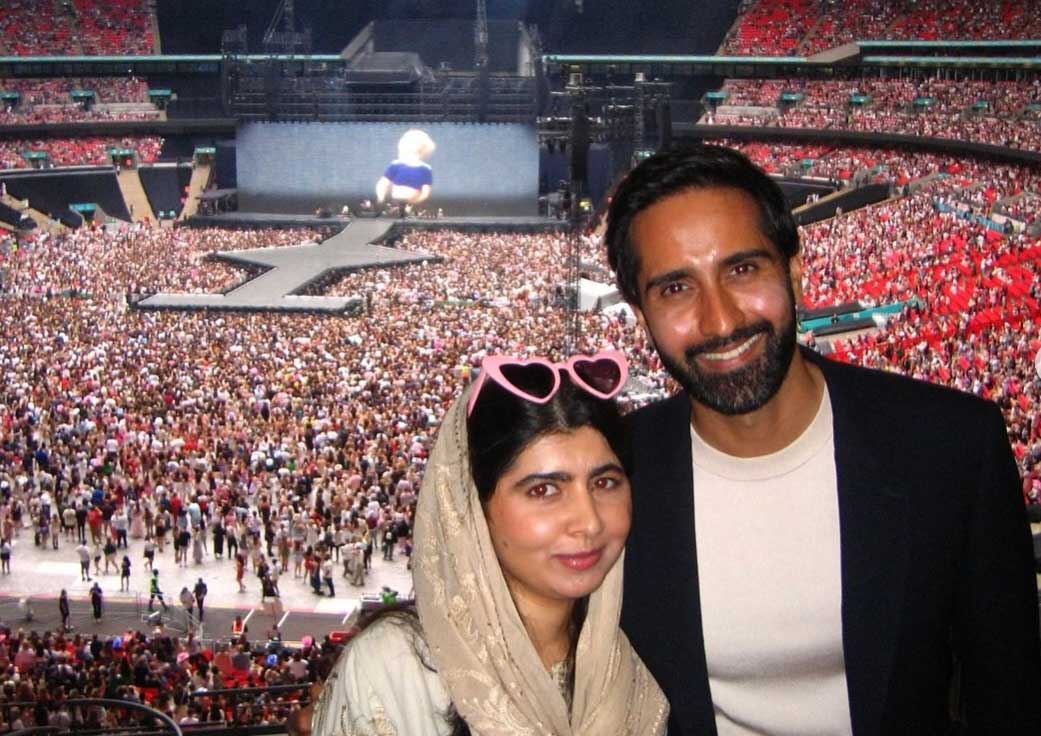 A woman with heart-shaped sunglasses on her head and a beige head covering stands next to a bearded man in a blazer and white T-shirt in a stadium full of people. Taylor Swift is on the large screen in the background.
