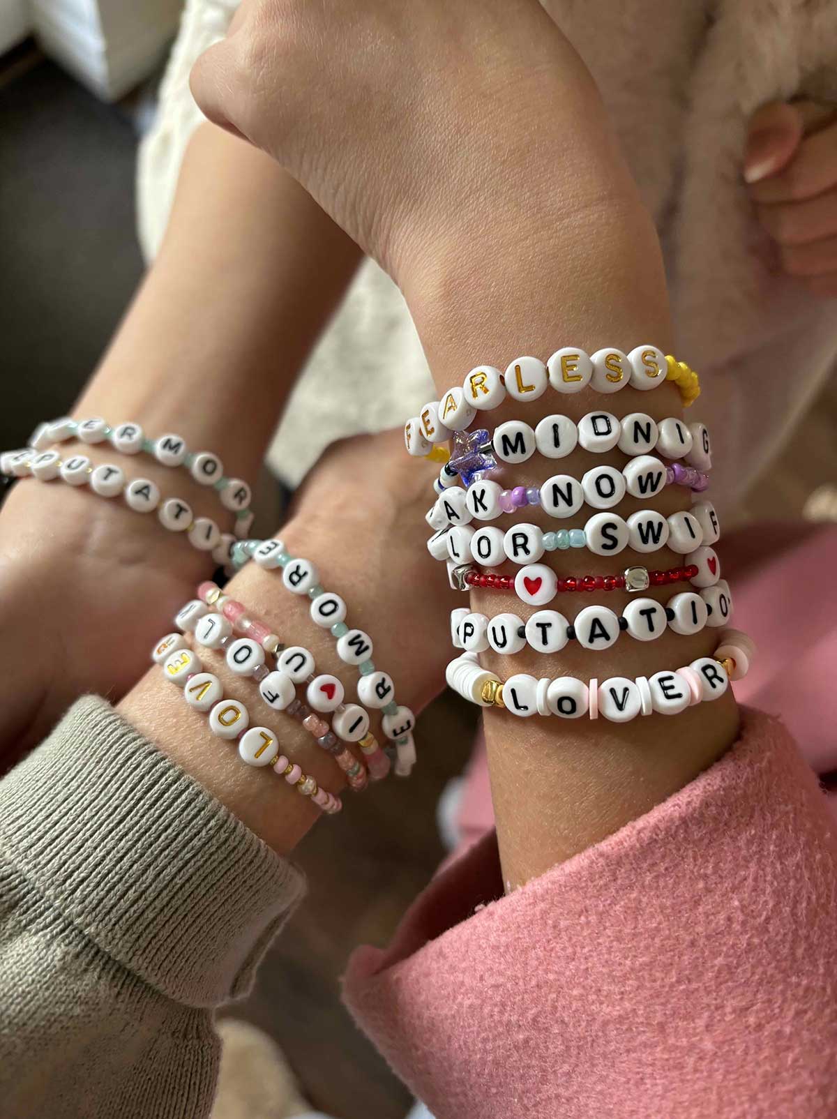 Young hands clasp each other to reveal wrists clad in stacks of friendship bracelets using small colourful plastic beads. Beads bearing letters of the alphabet spell the names of Taylor Swift albums and themes.