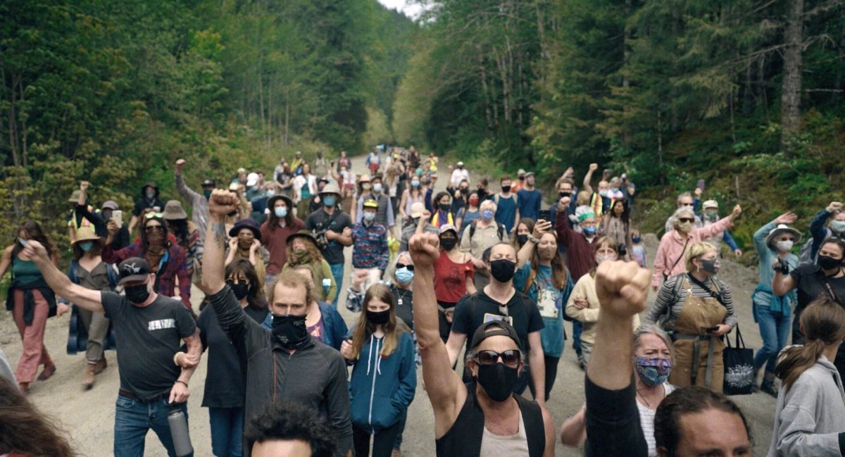 A group of protesters on a logging road wear cloth medical masks and raise their fists in the air.