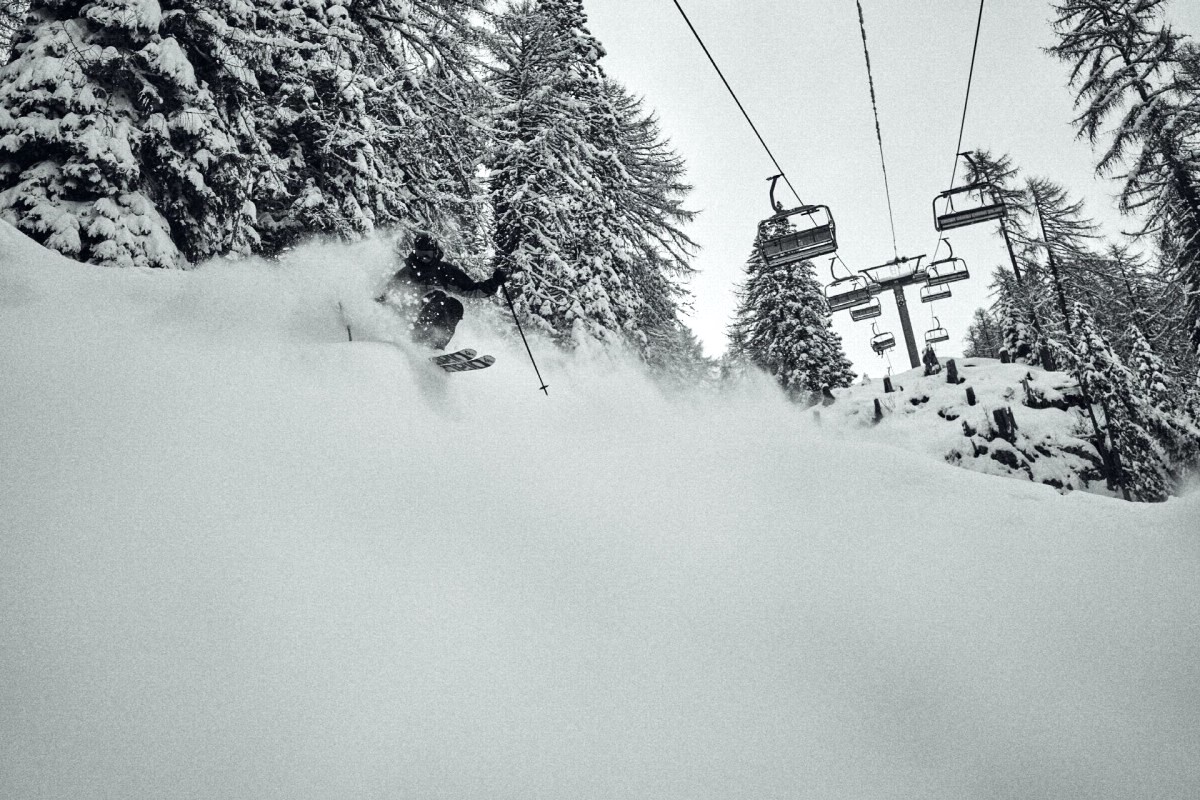 A skier in dark winter gear pushes through a powdery embankment under a chairlift.