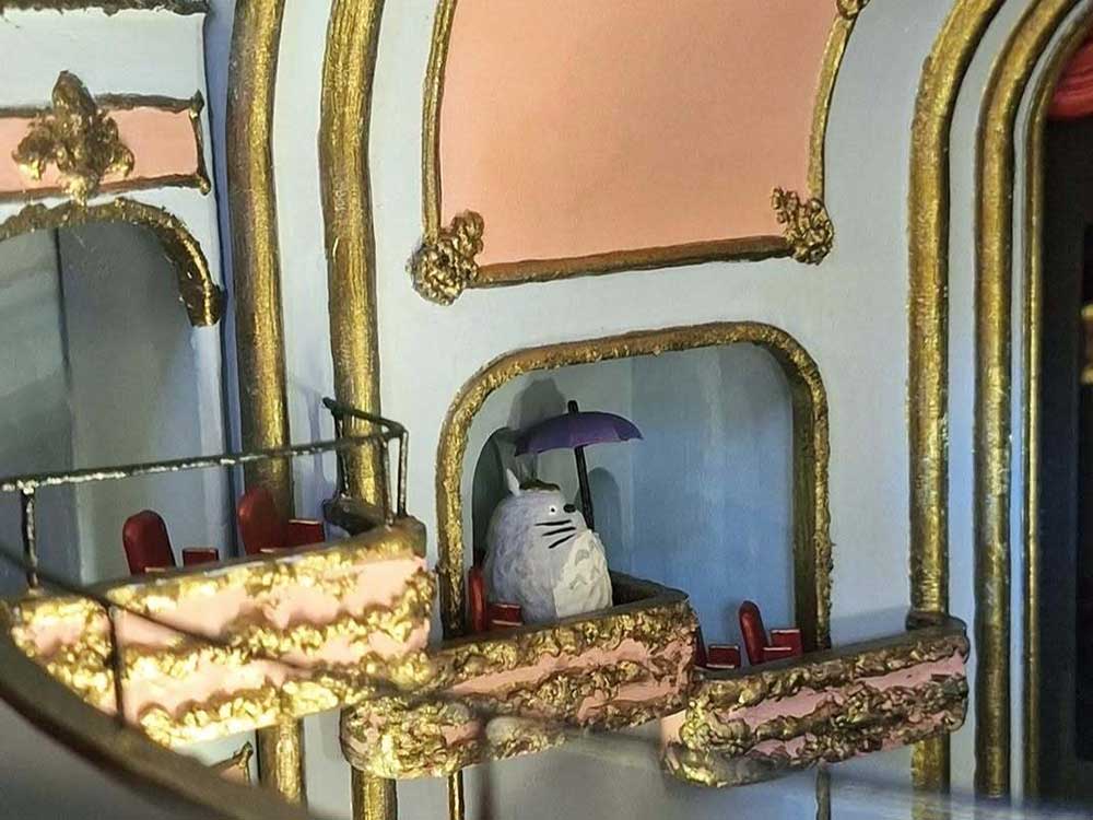 The interior of the seating area of a miniature theatre features gold trim, light pink accents, white walls and tiny red seats. A round grey cat-like creature holding a blue umbrella is in one of the balcony seats.