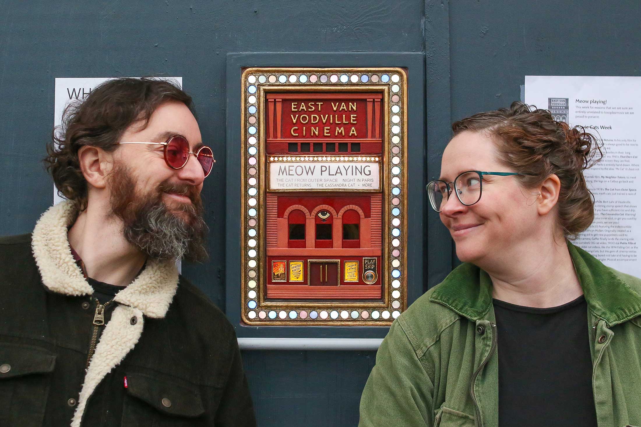 David Bynoe, left, and Janet Mader, right, stand on either side of a red rectangular ‘mini theatre’ surrounded by small lights. Its marquee reads “MEOW PLAYING” and above it are the words “East Van Vodeville Cinema.” Bynoe has dark wavy hair, light skin, round red sunglasses, a beard and a black jean jacket with a shearling collar. Mader has wavy reddish hair tied up in a messy bun, light skin and glasses. She wears an open green jacket over a black top.