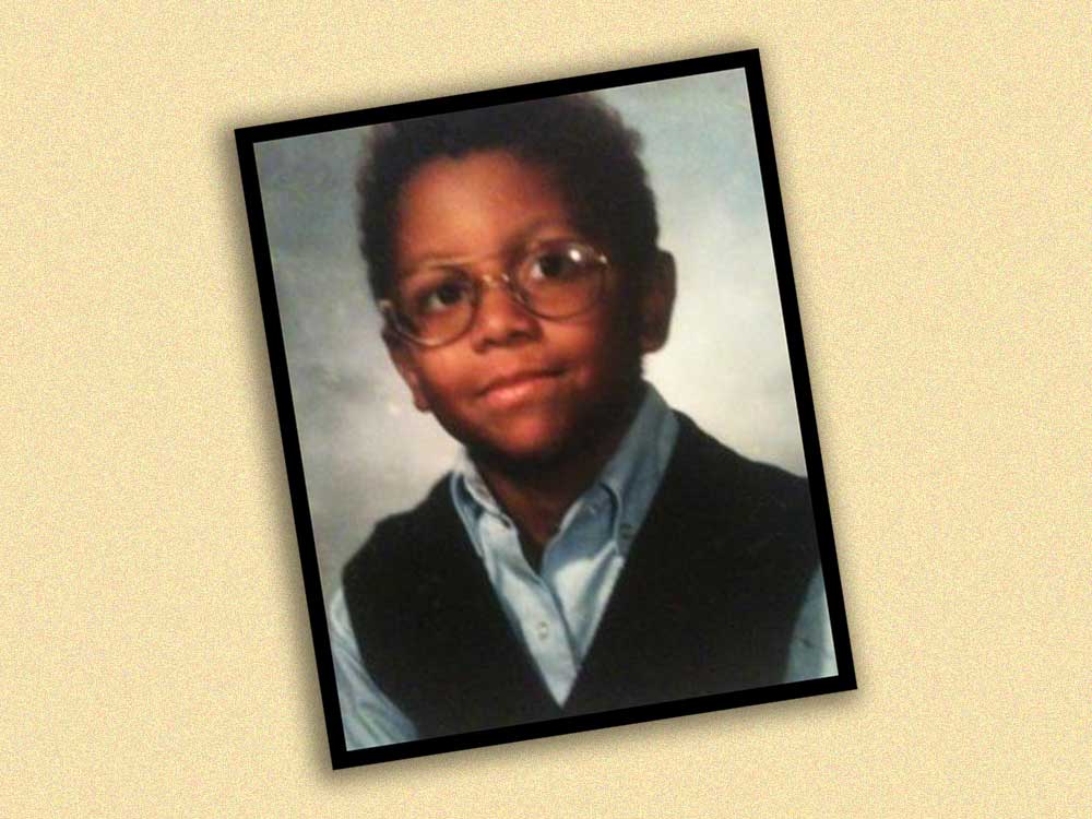 A wallet-sized portrait of a young boy in glasses, a dark blue vest and a light blue button-up shirt depicts the child smiling gently at the camera against a light blue background. 
