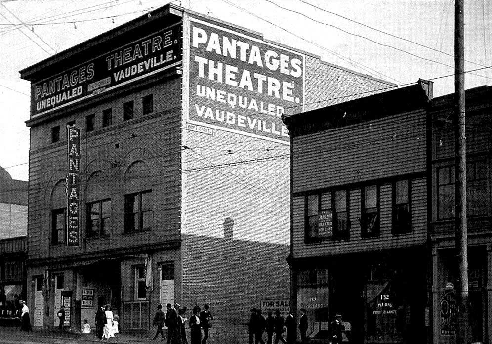 An archival black and white photo depicts a streetview of the Pantages Theatre in Vancouver, a tall brick building with the theatre’s name printed in large white text on the side of the wall.
