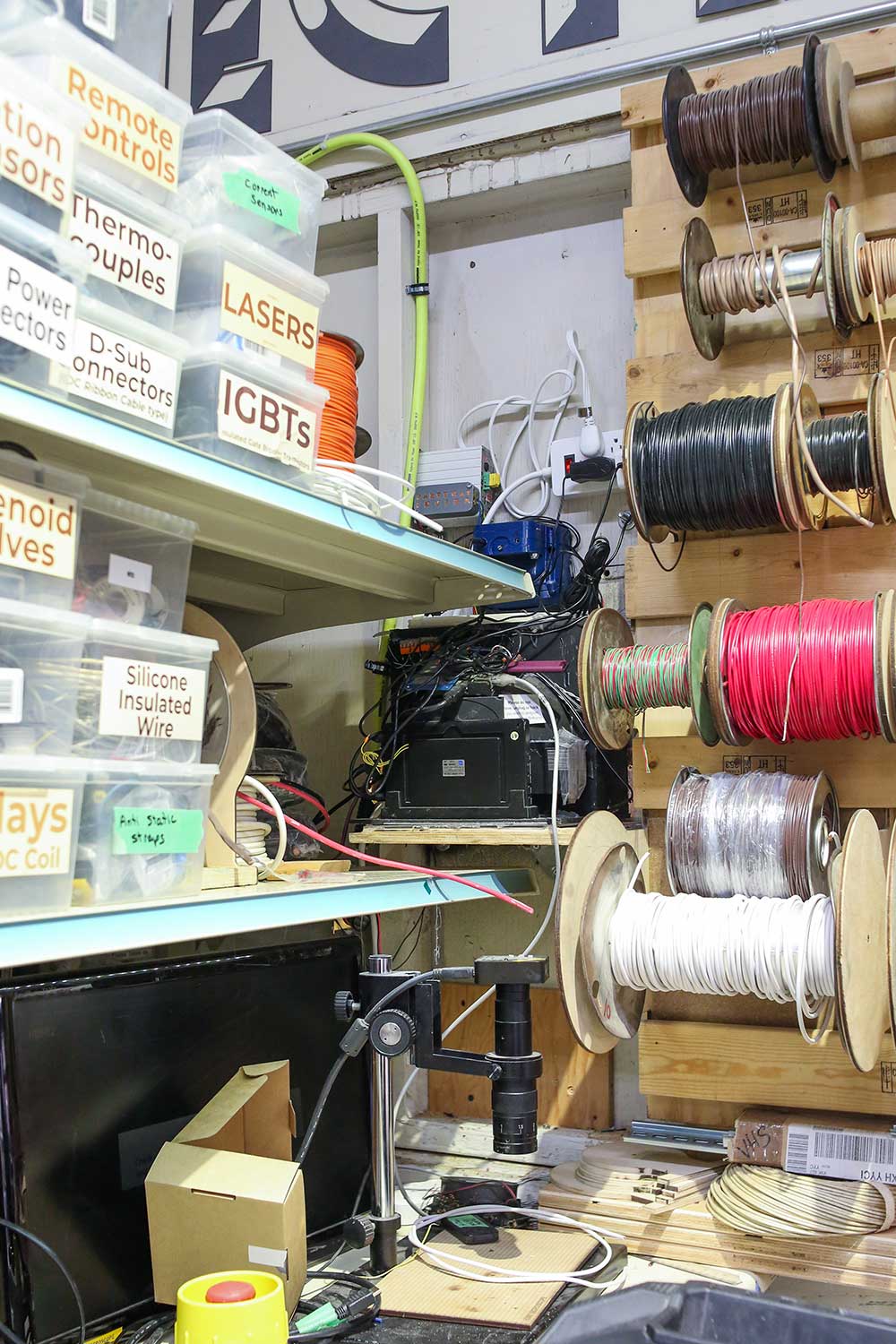 Colourful spools of coil are mounted on a plywood wall in an indoor studio space. To the left is a shelf featuring small clear plastic containers with labels like “thermo-couples” and “lasers.”