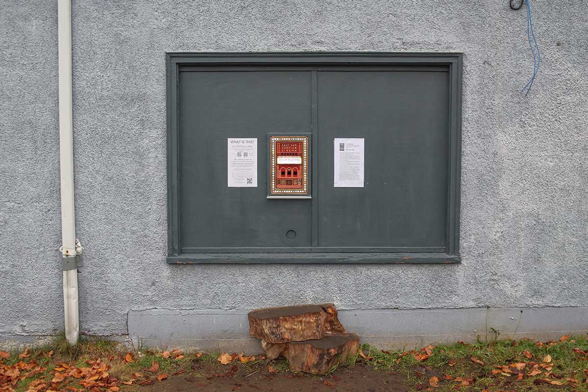 A large rectangular piece of wood is framed and painted dark grey against a light grey stucco wall. Below it is a makeshift stepping stool, above which stands a small red vertical rectangle, home of a mini cinema.