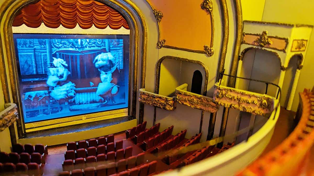 Top: On the lower left of the frame, a laptop displays a black and white digital illustration of the inside of the Pantages theatre; in the middle of the screen, a larger desktop computer screen displays a large sepia archival photograph of the theatre featuring a performer onstage and a live audience. Both monitors are surrounded by digital bric a brac and two coffee mugs. Middle: two miniature white side tables with pink drawers and gold handles sit on a tabletop against a piece of cardboard with red paint. A pair of hands holds a miniature gold balcony while another hand paints part of it white. Bottom: A look inside a miniature theatre on which a black and white animated film is playing on the movie screen. The seats are red and a grand red curtain stands above the screen.