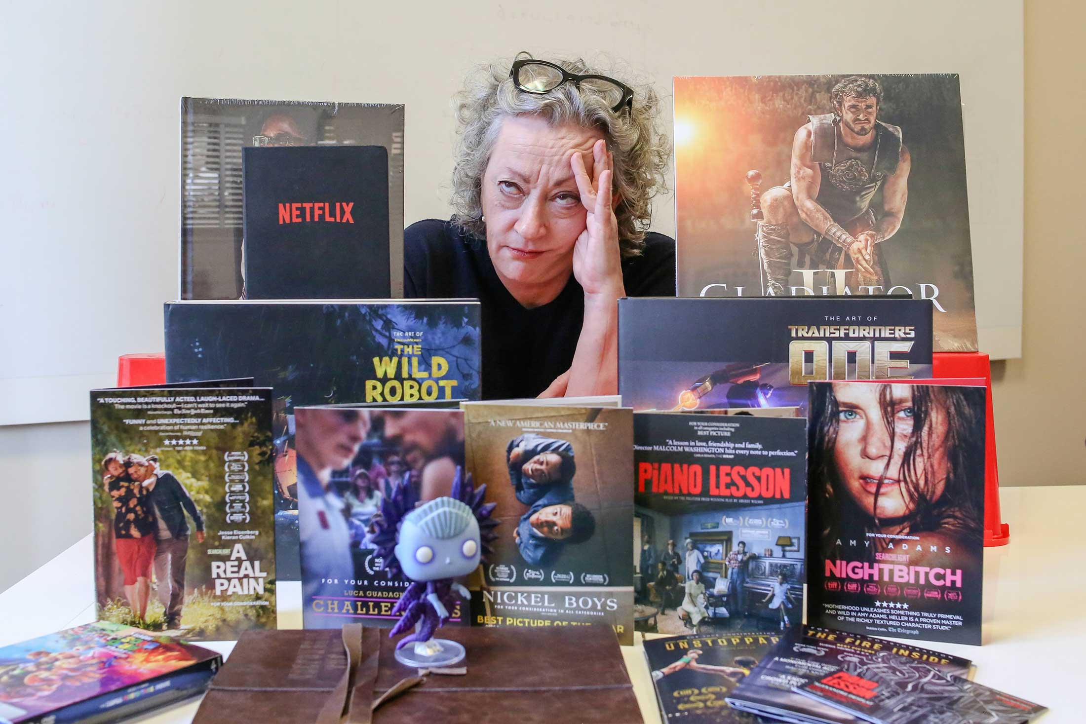 Dorothy Woodend has curly grey hair and is wearing a black top. She is seated at a table surrounded by movie studio merchandise. Her glasses are resting on top of her head and she rests her head in her hands with an exasperated expression. She is surrounded by movie-related swag including a vinyl record, DVDs, a book and a leather-bound script.