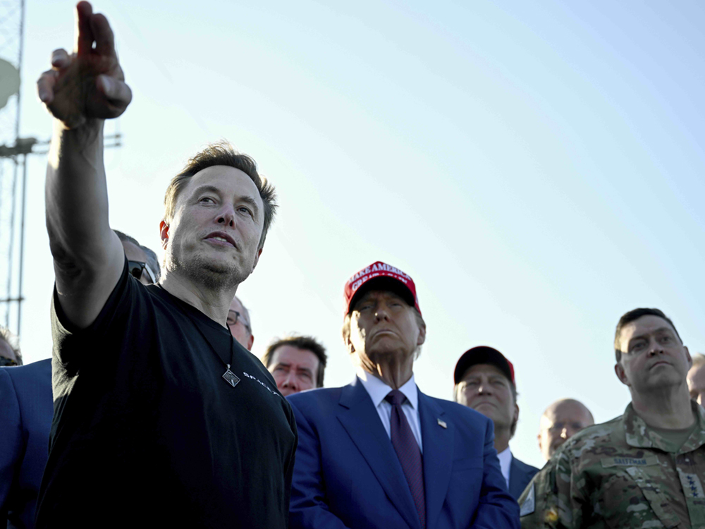 On the left, Elon Musk points upwards with two fingers. He is wearing a black T-shirt and has light brown hair and light skin. To the right is Donald Trump in a red 'Make America Great Again' ball cap and a blue suit over a white shirt and red tie. Around them is a group of men, one of them in military fatigues, under a light blue sky.