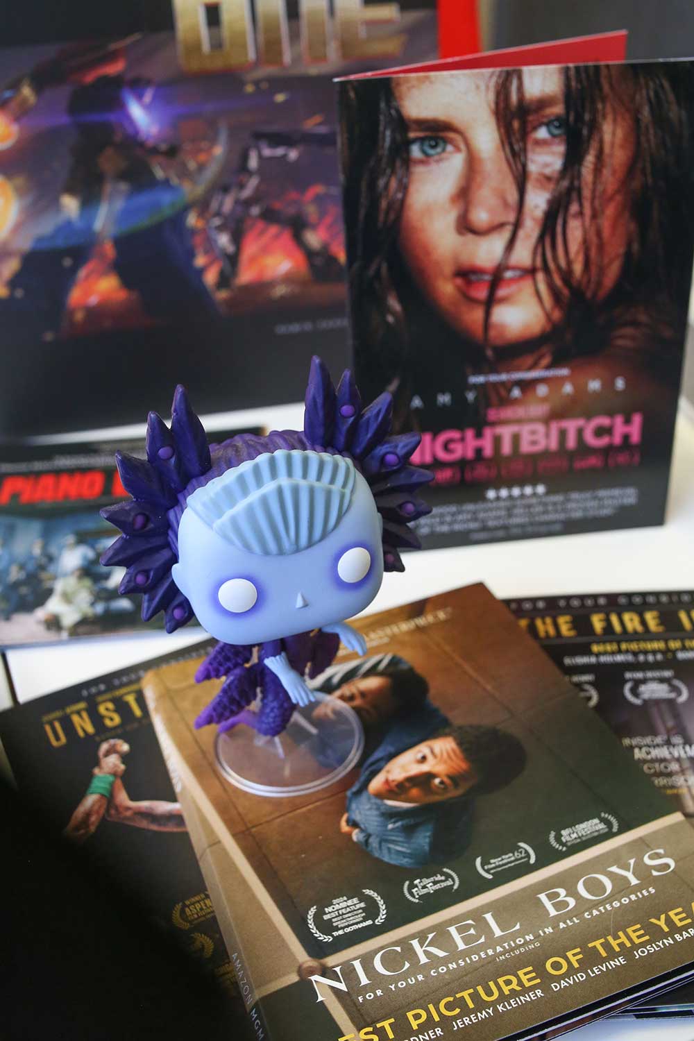 A small blue toy of a cartoon character with a light blue head and a spiky dark blue costume sits atop a DVD for the movie 'Nickel Boys.' In the background is press material for other movies, including 'Nightbitch.'